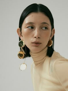 a woman with black hair wearing gold earrings and a beige turtle neck top, standing in front of a white wall