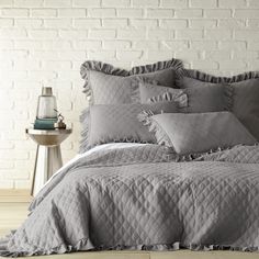 a bed with grey comforter and pillows on it next to a night stand in front of a white brick wall