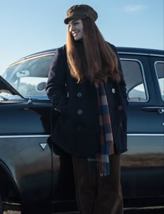 a woman standing in front of a car wearing a hat and coat with long red hair