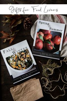 two cookbooks sitting on top of a wooden table next to some leaves and apples