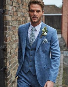 a man wearing a blue suit and flowered boutonniere stands in front of a brick wall