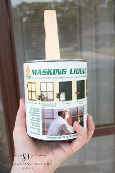 a person holding up a can of masking liquid in front of a window with a wooden stick sticking out of it