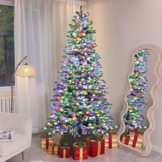 a brightly lit christmas tree in a living room next to a mirror with presents under it