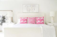 a white bed topped with pink pillows next to two lamps and a painting on the wall