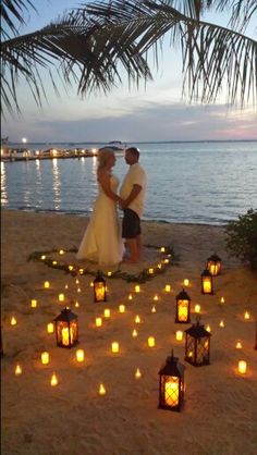 a bride and groom are standing on the beach surrounded by lit lanterns in the shape of hearts