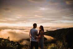 a man and woman standing on top of a mountain looking at the clouds in the distance