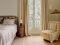 a bed sitting next to a window with curtains on it's sides and a chair in the foreground