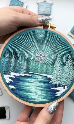 someone is holding up a cross stitch project with trees and water in the background, surrounded by spools of thread