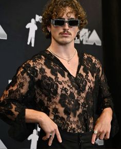 a man with curly hair wearing sunglasses pointing to the side while standing in front of a black background