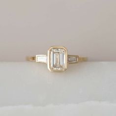 an emerald and baguette diamond engagement ring on a white surface with a light pink background