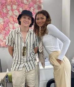 two people standing next to each other in front of a wall with flowers on it