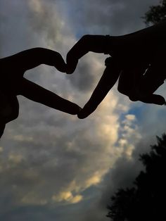 two hands in the shape of a heart against a cloudy sky with trees behind them