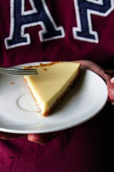 a person holding a plate with a piece of cheesecake on it and a fork in their hand