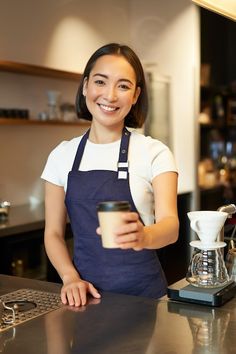 a woman holding a cup and smiling at the camera while standing in front of a coffee machine