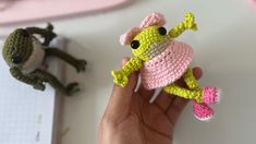 a hand holding a small crocheted frog doll next to two other stuffed animals