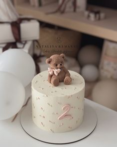 a small teddy bear sitting on top of a white cake with pink numbers and balloons in the background