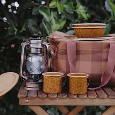 two mugs and a tea kettle sitting on a wooden table with a plaid bag next to it