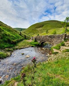 a small stone bridge over a stream in the middle of a green valley with hills behind it