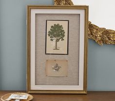 a framed picture with a tree on it next to a plate and gold leaf decoration