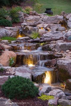 a small waterfall in the middle of a garden with rocks and plants around it, lit by lights