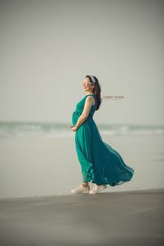 a pregnant woman in a green dress standing on the beach with her hands behind her back