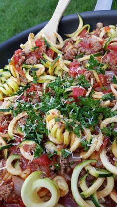 a skillet filled with pasta and vegetables on top of a green tablecloth next to a wooden spoon