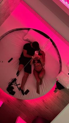 two people laying on a bed in a room with pink lights around them and one person holding a camera