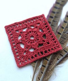 a red crocheted square sitting next to two feathers on top of a white surface