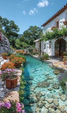 an outdoor garden with flowers, rocks and water in front of a white stucco house