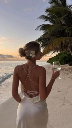 a woman standing on the beach with a glass of wine in her hand and palm trees behind her