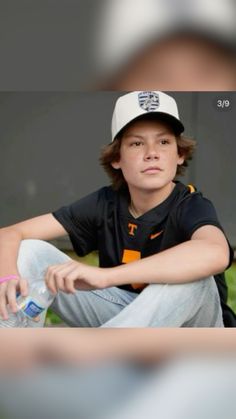 a young man sitting on the ground wearing a baseball cap and holding a water bottle