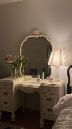 a white desk with a mirror, lamp and flowers on it next to a bed