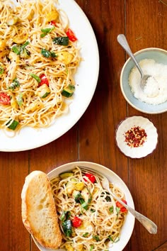 two plates of pasta and bread on a wooden table with bowls of sauces next to them