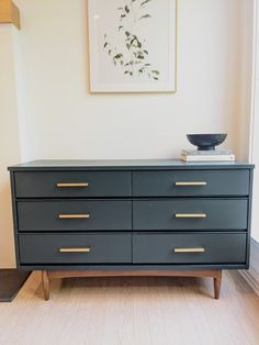 a black dresser with gold handles in a white walled room next to a painting on the wall