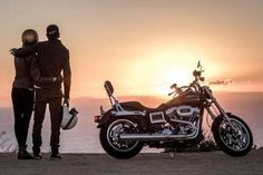 two people standing next to a motorcycle on top of a hill with the sun setting in the background