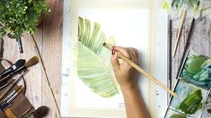 a person is holding a paintbrush and painting a leaf on a piece of paper