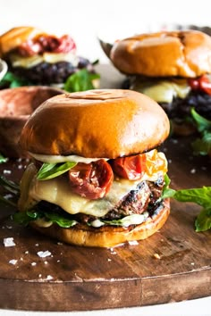 two burgers sitting on top of a wooden cutting board next to buns filled with meat and vegetables