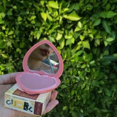 a person holding up a pink compact mirror in front of a green bush and shrubbery