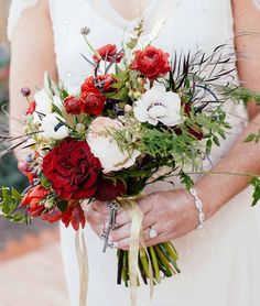 a woman in a white dress holding a red and white bouquet with greenery on it