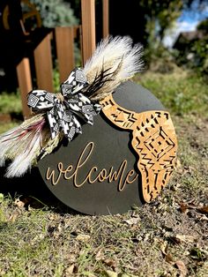 a welcome sign with feathers on it sitting in the grass next to a wooden fence