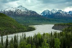 the mountains are covered in snow and green trees, with a river running between them