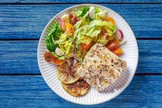 a white plate topped with meat and salad on top of a blue wooden table next to an orange slice