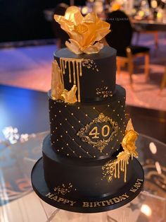 a black and gold birthday cake with flowers on it's tiers is sitting on a glass table