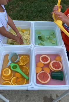 two children are playing with their food in trays on the grass, and one child is holding a yellow plastic spoon