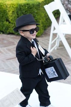 a little boy dressed up in a suit and tie carrying a briefcase while wearing sunglasses