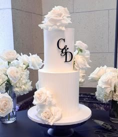 a three tiered white wedding cake with monogram on top and flowers in vases