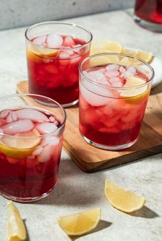 two glasses filled with red liquid and lemon slices on a cutting board next to sliced lemons