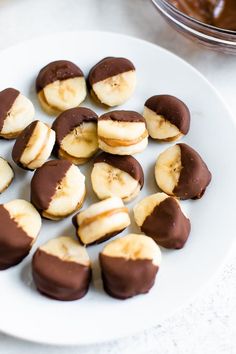 chocolate covered bananas on a white plate next to a bowl of peanut butter and banana slices