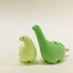 two small plastic dinosaurs sitting next to each other on a white surface, one green and the other light green