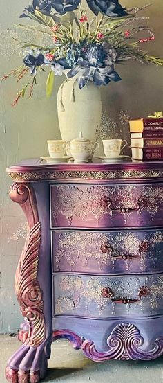 a purple dresser with flowers in a vase on top and books sitting on the bottom